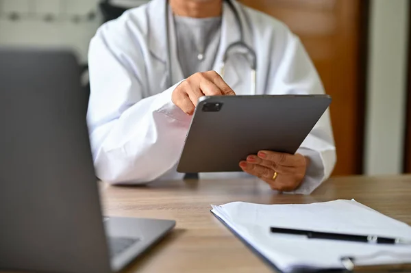 cropped image, Professional female doctor in white gown sits at her desk using digital tablet to check her surgery schedule.