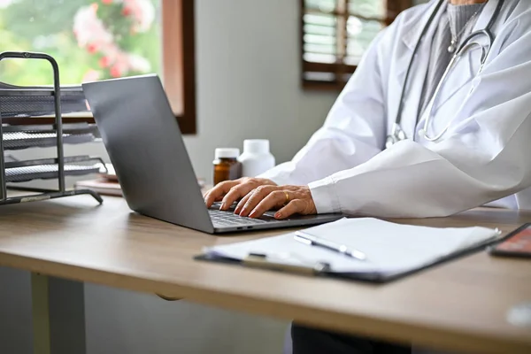 Female doctor in white gown using laptop computer, typing on keyboard, researching medical online paper on internet, checking her schedule, working in the office. cropped image