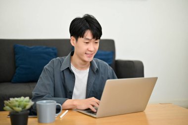 Akıllı Asyalı genç adam dizüstü bilgisayar kullanıyor, klavyede yazıyor, internette bir şeylere bakıyor, oturma odasında çalışıyor..