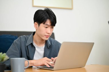 Odaklanmış ve yakışıklı Asyalı adam dizüstü bilgisayar kullanıyor, online proje yapıyor, oturma odasında çalışıyor..