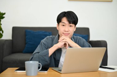 Gülümseyen ve yakışıklı Asyalı genç adam asgari oturma odasında oturuyor, elleri çenesinde, kameraya bakıyor, dizüstü bilgisayar sehpanın üzerinde..