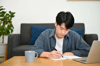 Odaklanmış genç Asyalı üniversite öğrencisi günlük yazıyor ya da bir okul kitabına not alıyor, oturma odasında ödev yapıyor..