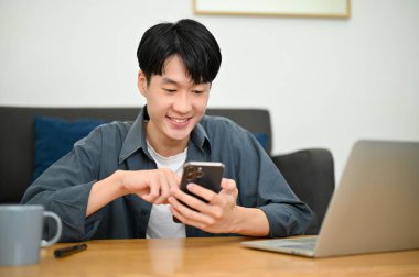 Neşeli ve yakışıklı Asyalı genç adam akıllı telefonunda mobil uygulama kullanıyor, sosyal medya platformunda kayıyor, oturma odasında dinlenirken arkadaşlarıyla sohbet ediyor..