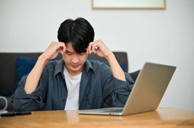 Düşünceli ve stresli genç Asyalı erkek üniversite öğrencisi okul projesini düşünüyor, oturma odasında oturuyor..