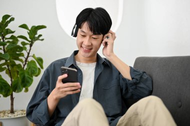 Rahat ve mutlu Asyalı adam, kablosuz kulaklık takıyor, müzik dinliyor, oturma odasındaki kanepede dinlenirken telefonda müzik listesi yapıyor..