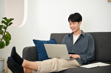 Uzaktan kumandalı rahat ve soğuk Asyalı adam oturma odasındaki kanepede otururken dizüstü bilgisayar kullanıyor..