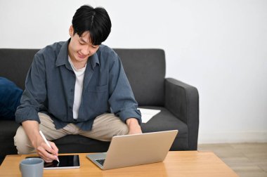 Yakışıklı Asyalı genç adam portatif dijital tabletini kullanıyor, oturma odasında çalışıyor..