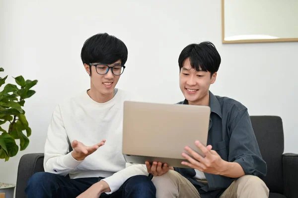Two of young Asian male best friends are watching a sports match on a laptop in the living room together.