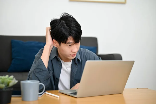 Confused young Asian man experiencing internet connection issues at work, getting some trouble on his laptop, pensively thinking about his project.