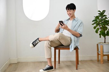 Hoş, yakışıklı, sıradan giyimli Asyalı bir adam minimumda oturma odasında akıllı telefonunu kullanarak bir sandalyeye oturuyor..