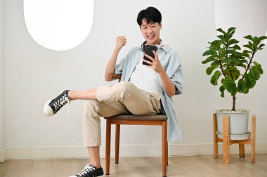 Heyecanlı ve neşeli Asyalı genç adam yumruğunu sıkmış, akıllı telefon ekranına bakıyor, kutlama, galibiyet, zafer, iyi haber.