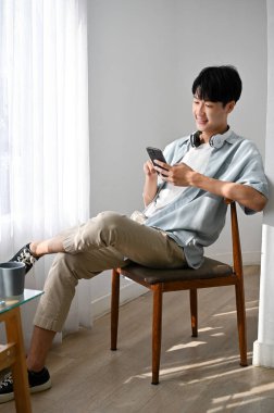 Yakışıklı Asyalı bir adam akıllı telefon kullanıyor, arkadaşlarıyla sohbet ediyor, sandalyede otururken mobil uygulama kullanıyor..