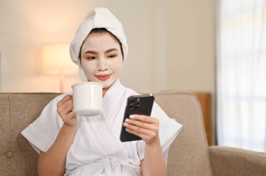 Çekici genç Asyalı kadın bornozlu ve yüzünde kilden bir maskeyle, kahvesini yudumluyor ve oturma odasındaki koltukta dinlenirken telefonunu kullanıyor..