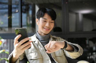 Gülümseyen ve mutlu bin yıllık Asyalı adam telefonunu tutuyor, kafeteryada dinlenirken akıllı saatini kontrol ediyor..