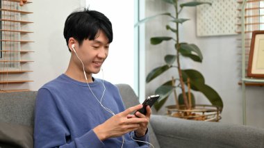 Mutlu ve yakışıklı Asyalı genç adam telefonuyla oturma odasında kulaklıklarıyla müzik dinliyor..