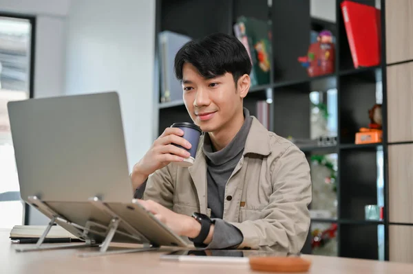 Attractive millennial Asian male businessman in casual clothes focusing on his task, using laptop computer and sipping coffee.
