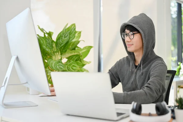 Smart and nerd young Asian male website developer or programmer wearing eyeglasses and hoodie sweater focusing on his task on computer in the office. side view