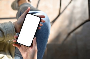 Top view, A female holding a smartphone white screen mockup over blurred background. A female scrolling on social media while relaxing at the cafe.