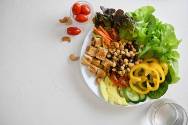 Top view, Buddha bowl mixed vegetables with grilled tofu on a white table with a glass of water. Vegan food and plant based concept