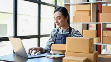 Charming and attractive millennial Asian female online shop owner using laptop computer, happy with her sale orders, working in her home office.