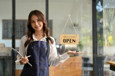 Beautiful and friendly young Asian female waitress or coffee shop staff, in an apron with her tablet, stands at the entrance door of her cafe, welcoming customers.