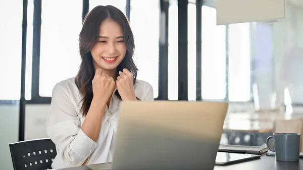 Cheerful and happy young Asian female office worker looking at laptop screen, receiving great news, getting job promotion or project approved.