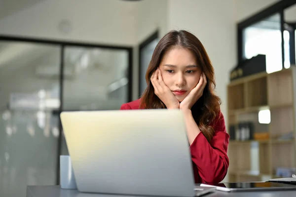 Unhappy and bored young Asian businesswoman sits at her office desk looking at laptop screen, feeling tired from work, no work passion. upset, overwork, thoughtful, depressed.