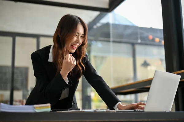 Cheerful and happy young Asian female office worker leaning on table, showing clenched fits, looking at laptop screen, receiving great news, getting job promotion or project approved.