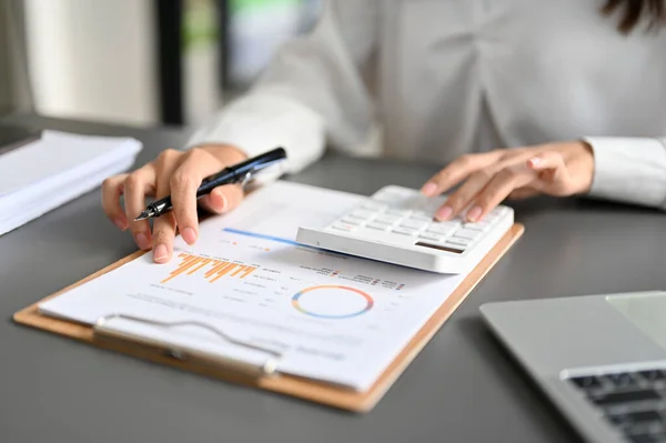 Professional female accountant or financial worker using calculator, calculating business sales accounts, reviewing financial report. cropped and close-up image