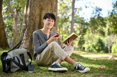Handsome and happy young Asian male college student sits under the tree in the park, eating apple while reading a book. leisure and hobby concept