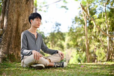 Handsome and relaxed young Asian man in casual clothes meditating under the tree in the greenery park.