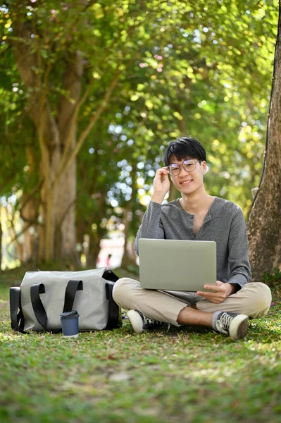 Portrait, Smart and happy young Asian male college student wearing glasses, sits under the tree in the campus park with his portable laptop computer. Uni student lifestyle concept