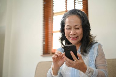 Charming and happy 60s aged Asian woman using her smartphone, chatting with her friends while relaxing in her living room. Retirement lifestyle concept