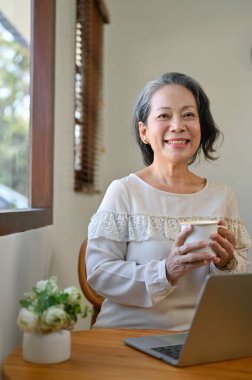 Portrait, Charming 60s retired Asian woman holding a tea cup, smiling and looking at the camera while relaxing in the cafe.