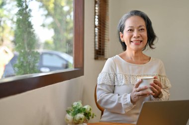 Charming and happy 60s retired Asian woman holding a tea cup, smiling and looking at the camera while relaxing in the cafe.