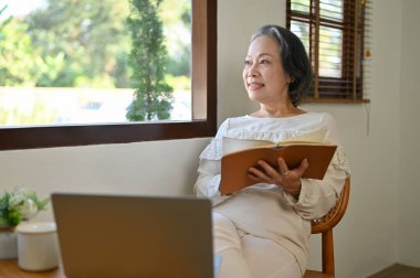Relaxed and calm 60s retired Asian woman looking out the window, daydreaming while reading a book in her home living room. lifestyle concept