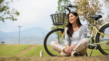 Relaxed and happy young Asian woman in casual clothes sits on the grass, taking a rest after ride a bike in beautiful nature park on the morning. outdoor activity concept