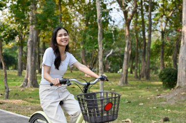 Joyful and happy young Asian woman riding a bicycle in the public park on the weekend, enjoys doing outdoor activity.