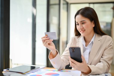 Charming young Asian businesswoman sits at her desk, using her mobile banking to pay an online shopping bills. cashless payment concept