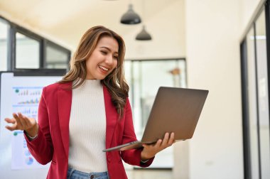 Professional and excited millennial Asian businesswoman stands in her office using portable laptop computer, happy with project's results.