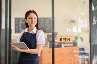 Charming young Asian female barista or waitress in apron stands in front of the coffee shop's entrance door with her portable tablet. small business concept
