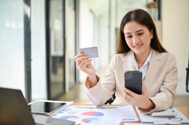 Happy and charming millennial Asian businesswoman or female boss using mobile banking to pay an online bill at her desk. internet banking, mobile banking, credit card online payment.