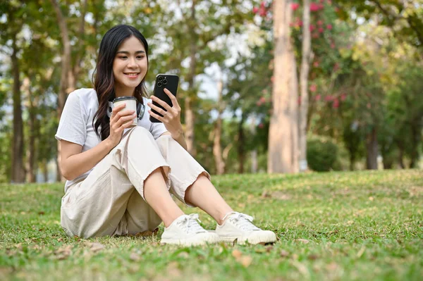 Charming young Asian woman sits on grass, sipping coffee and using her phone while relaxing in the greenery park. hobby and leisure concept