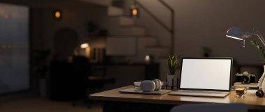 Modern home workspace at night with laptop white screen mockup, light from table lamp, headphone and accessories on table over blurred background of darkness room. 3d render, 3d illustration