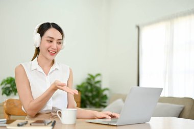 Attractive and cheerful millennial Asian woman or businesswoman wearing wireless headphones, having an online meeting or webinar via laptop. work from home concept