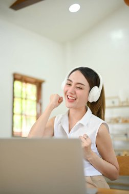 Portrait, Beautiful and overjoyed millennial Asian woman enjoys listening to music through her headphones, looking at laptop screen, dancing at her desk.