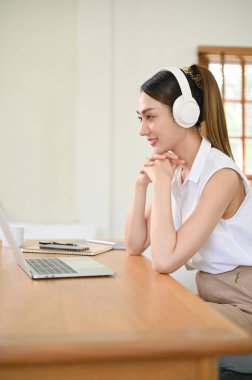 Gorgeous and attractive millennial Asian woman sits at her desk hands on chin, wearing headphones, listening to music or joining in business online meeting. side view
