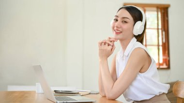 Attractive and charming millennial Asian woman wearing headphones, listening to music while sitting at her desk with her laptop and stationery. side view