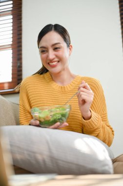 Happy and attractive millennial Asian woman in casual comfy clothes eating salad bowl while enjoys watching movie via laptop on sofa in her living room. lifestyle concept
