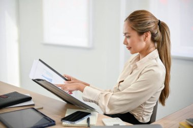 side view, Professional and attractive millennial Asian businesswoman or female manager examining business financial report on a folder binder at her desk